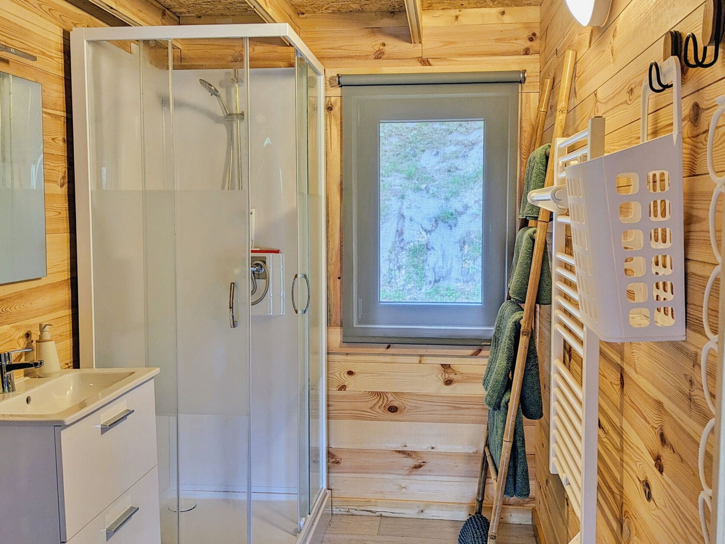 Une salle de bain rustique, avec des murs en bois, une douche en verre, un lavabo et une fenêtre donnant sur la forêt.