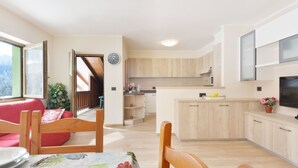 Eine moderne Küche mit Holzschränken, ein Essbereich mit Tisch für zwei Personen und ein großes Fenster mit Blick ins Freie.