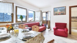 A dining area with a table set for four, a red armchair, and a view of snowy mountains through large windows. - Bernard by Interhome (Campitello)