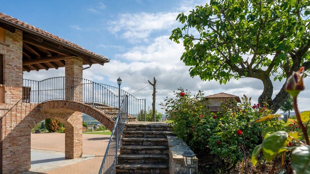 A stone staircase leading to a covered area with a metal railing, surrounded by greenery and a brick building with a tiled roof.