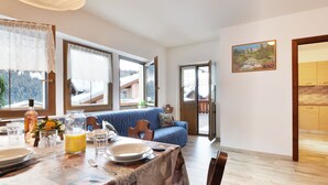 A dining area with a wooden table set for two, a blue sofa, and a view of snowy mountains through large windows. - Bernard by Interhome (Campitello)