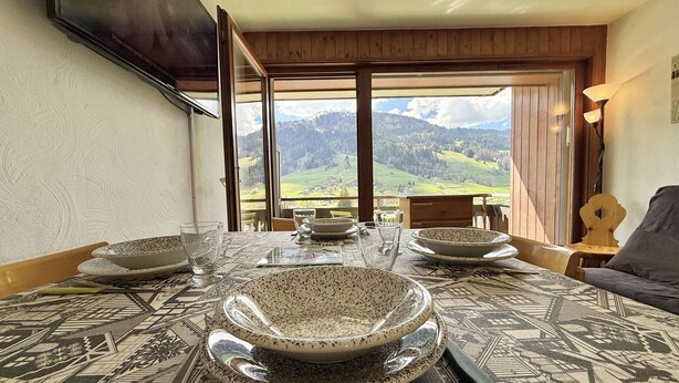 A dining table set with plates, glasses, and cutlery, with a scenic mountain view through large windows. - Cozy 2-Room Cabin Apartment in Praz Sur Arly, Close to Ski Lifts, Sleeps 5, Parking Included (Praz-sur-Arly)