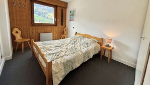 A bedroom with a wooden bed, a chair, a small table with a lamp, a window with a view of a green landscape, and a wooden wall with a 'LOVE' sign. - Cozy 2-Room Cabin Apartment in Praz Sur Arly, Close to Ski Lifts, Sleeps 5, Parking Included (Praz-sur-Arly)