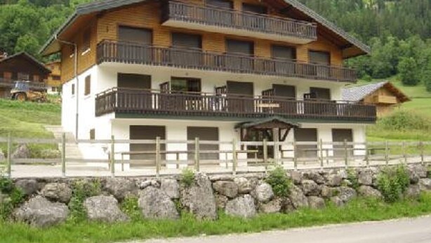 A multi-story wooden building with balconies, set in a mountainous area with greenery.