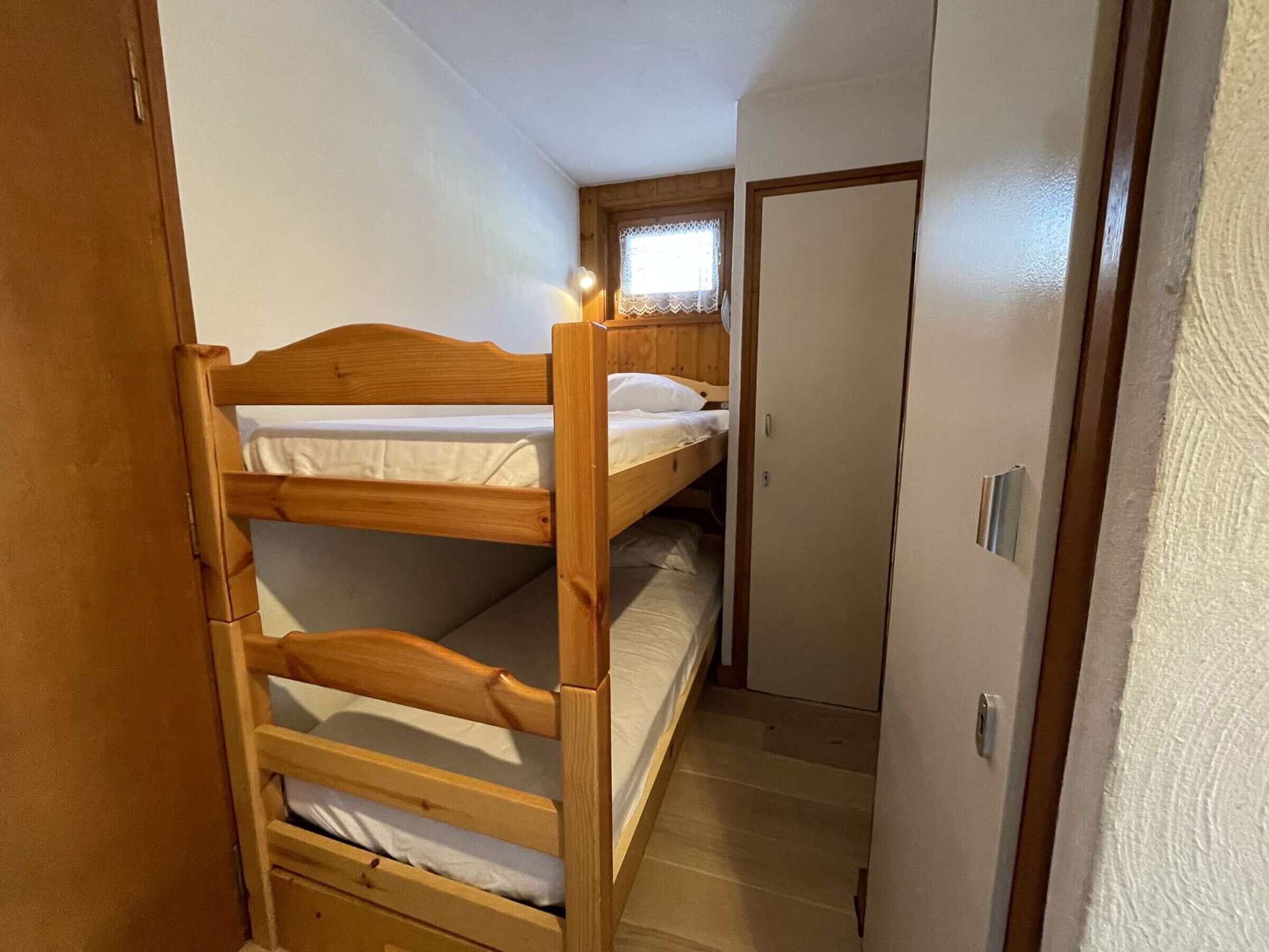 Habitación con literas, cabecera de madera, una ventana pequeña con cortinas de encaje y una puerta que da a otra habitación.