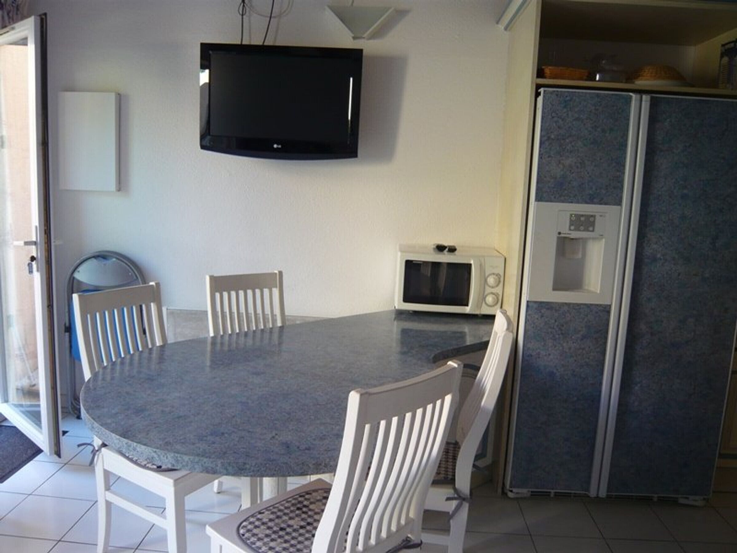 A dining area with a round table, four white chairs, a flat-screen TV mounted on the wall, a microwave, and a refrigerator with a water dispenser.