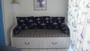 Una habitación con sofá cama con cojines a rayas, un cuadro enmarcados con flores, una lámpara sobre un estante azul y cortinas con motivos náuticos.