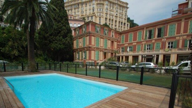 A swimming pool with a wooden deck, surrounded by a fence, and a building with red and green shutters in the background. - Charming 1-BR Apt in Central Menton with Pool, Terrace & Parking (Menton)