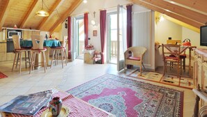 Ein geräumiges Dachzimmer mit Holzbalken, einem Essbereich mit Stühlen und Tisch, einer Küchenzeile mit Mikrowelle und einem großen Fenster mit Vorhängen.