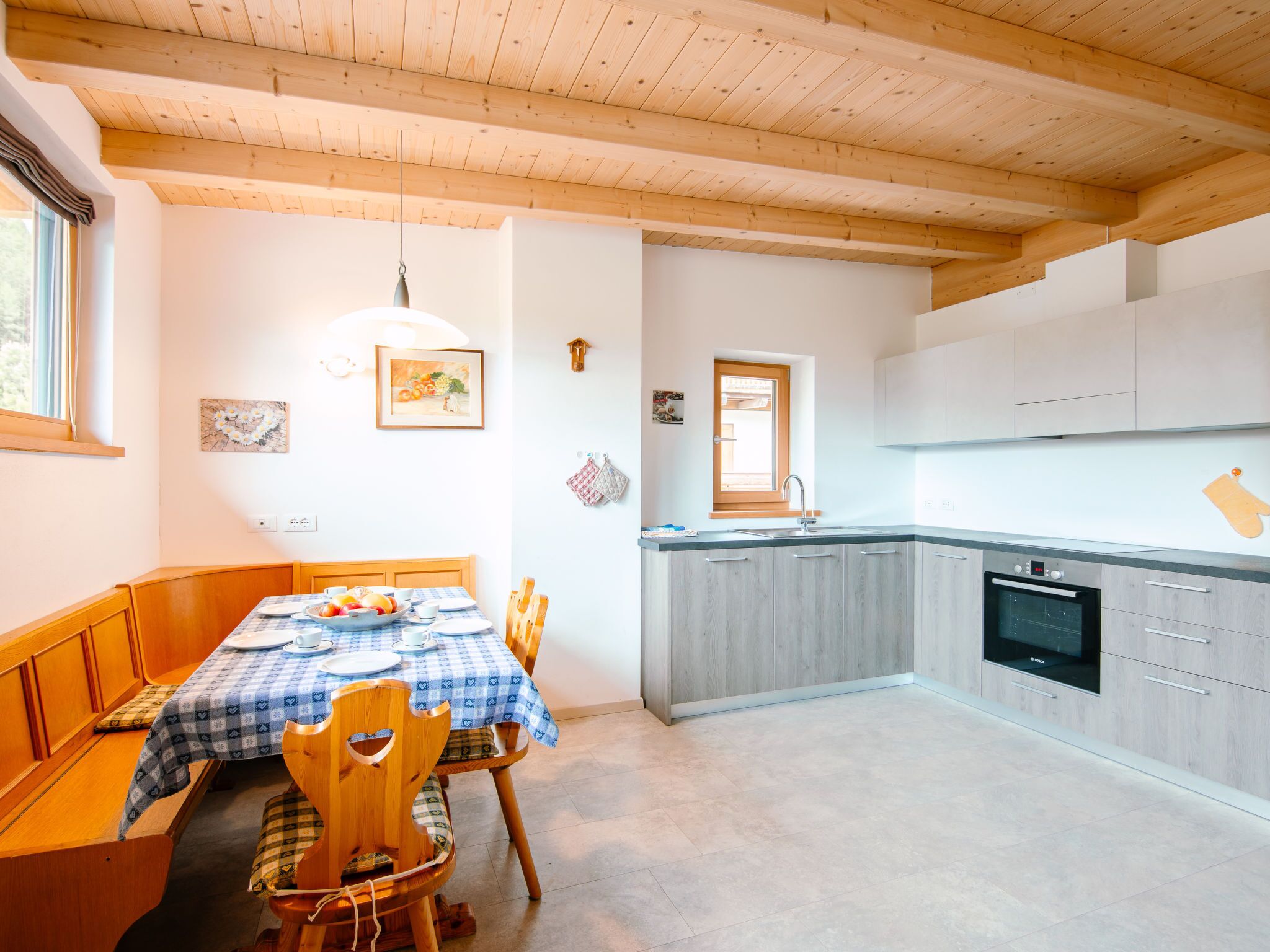 A dining area with a wooden table and chairs, a kitchen with grey cabinets and a built-in oven, and a window with a view of greenery.