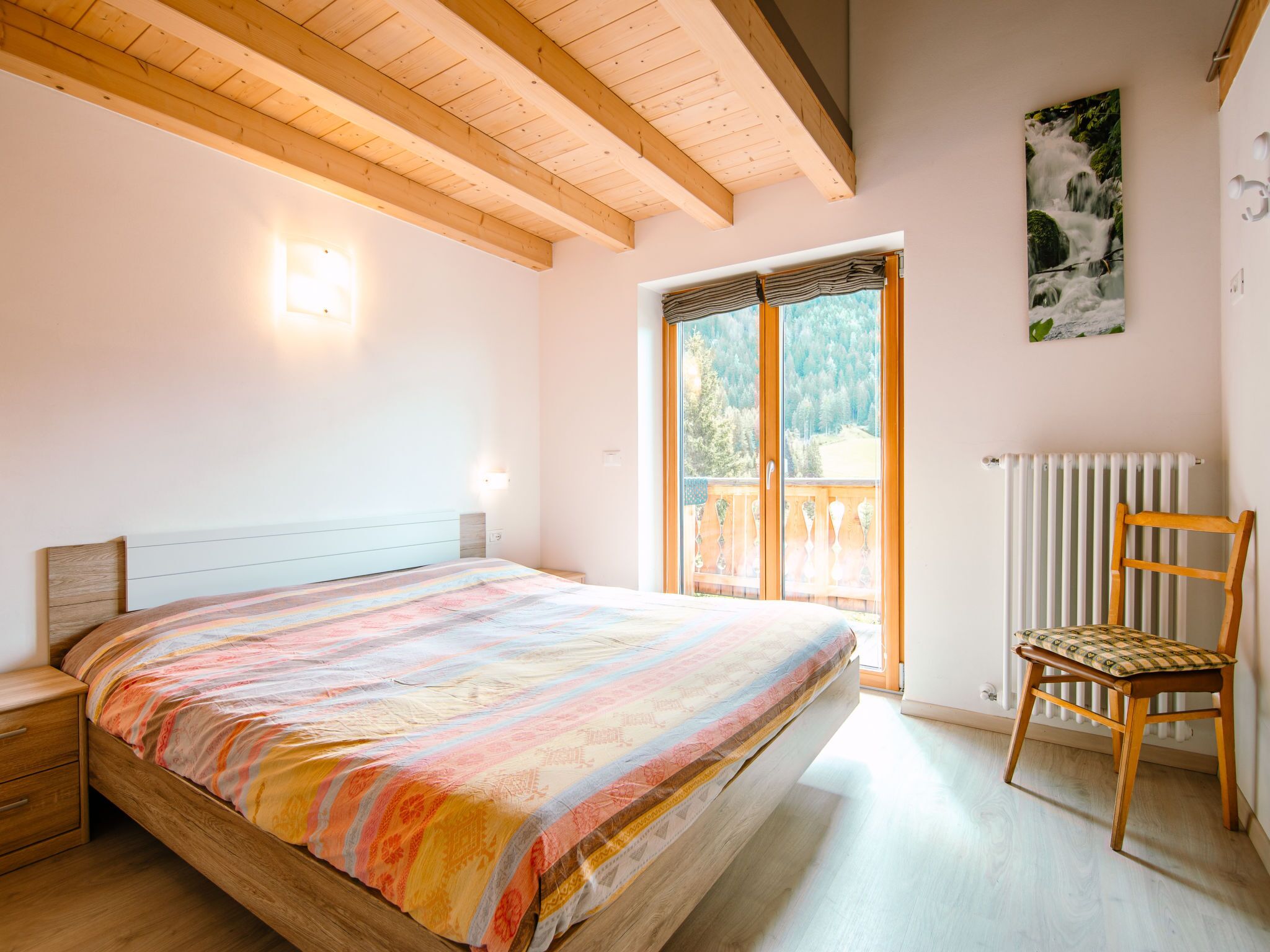 Een slaapkamer met een bed, een houten stoel, een radiator en een raam met uitzicht op bomen.