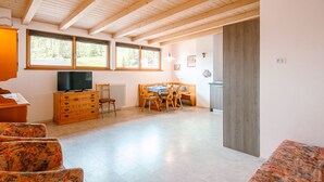 Una habitación amplia con vigas de madera en el techo, un televisor sobre un mueble de madera, una zona de comedor con mesa y sillas, y ventanas grandes que ofrecen vistas al exterior.