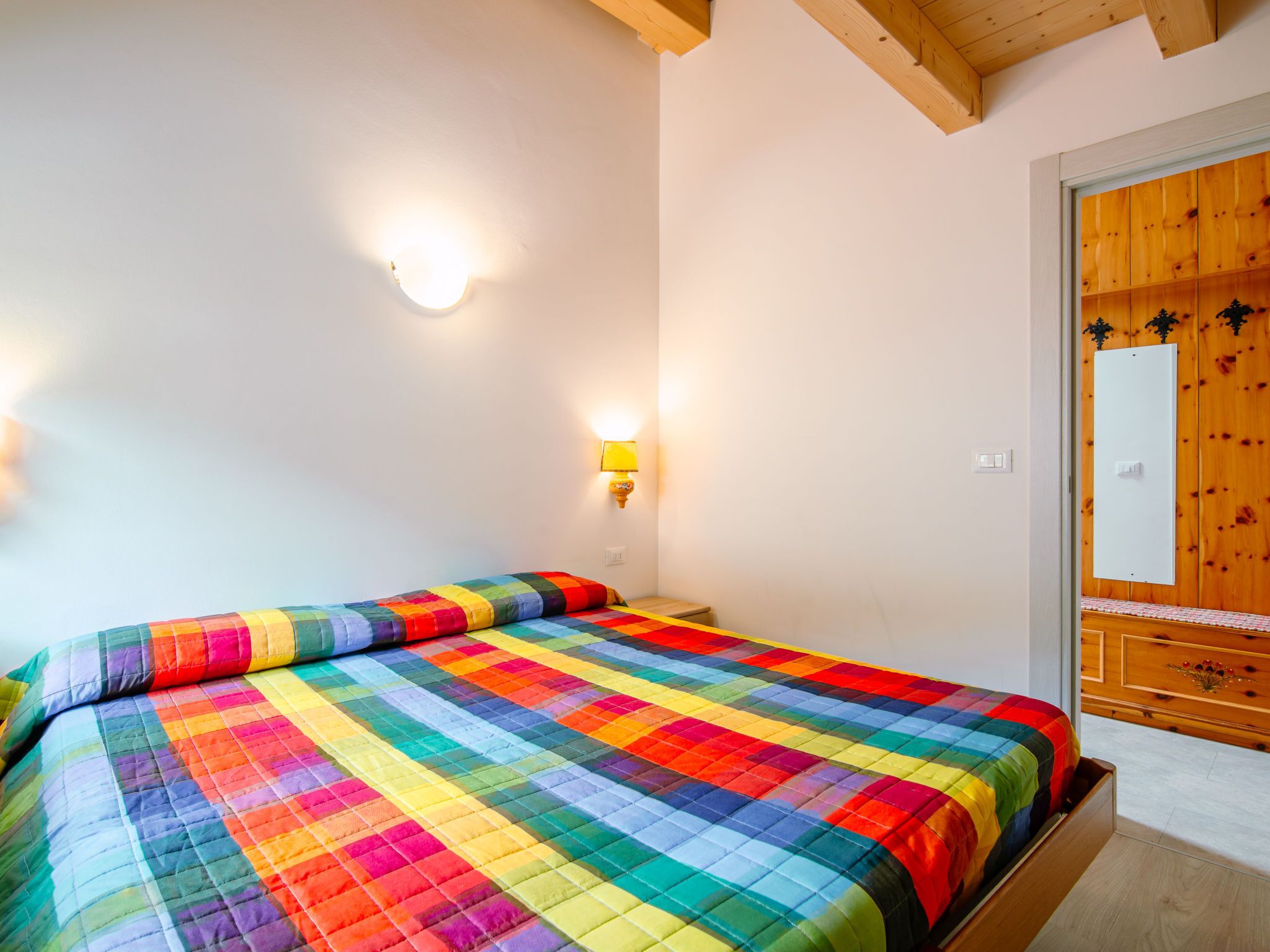 Een slaapkamer met een kleurrijk geborduurd bedovertrek, een houten deur met een spiegel, en een wandlamp.