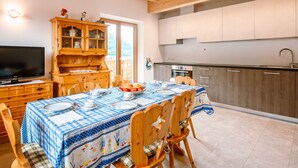 Un comedor con una mesa dispuesta para cuatro, un aparador de madera con puertas de vidrio, un televisor y una zona de cocina con gabinetes grises y un lavamanos de acero inoxidable.