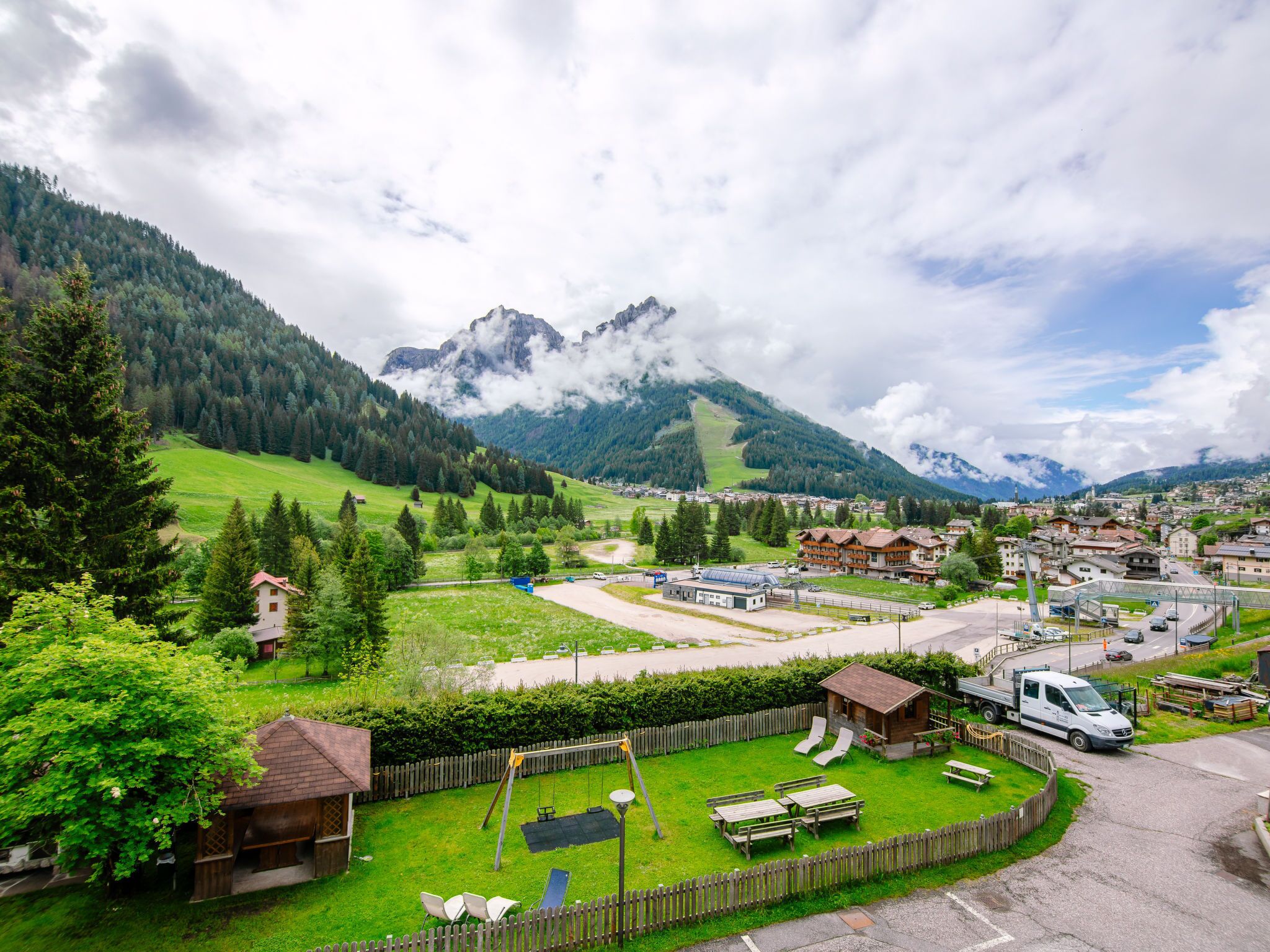 Un piccolo borgo rurale con un'area giochi, tavoli da picnic e un furgone parcheggiato vicino alla strada.