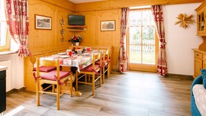 A cozy dining room with a wooden table set for four, surrounded by chairs with red cushions. A television is mounted on the wall above the table.