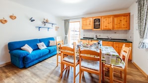 Una sala de estar acogedora con un sofá azul y una mesa de comedor de madera, una cocina con armarios de madera y un ventanal con cortinas.