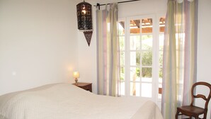 Un dormitorio con una cama grande, una silla de madera, una mesita de noche con lámpara y una ventana con cortinas.