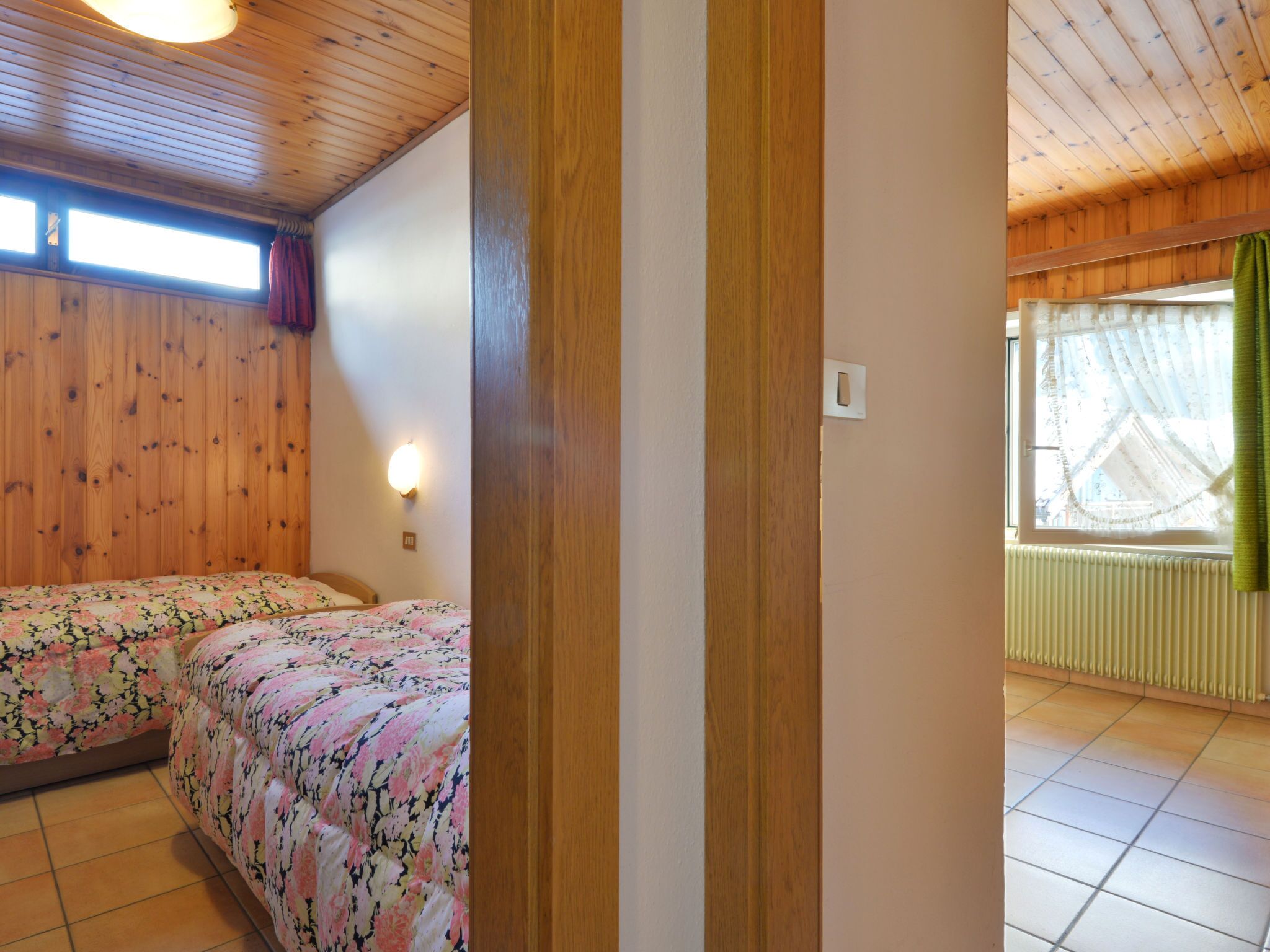 Une pièce aux murs recouverts de lambris en bois, un lit avec une literie à motifs floraux, une fenêtre avec des rideaux et un radiateur.