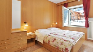 Une chambre avec des murs en bois, un lit recouvert d’une literie à motifs floraux, une table de chevet, une commode avec des tiroirs et une fenêtre avec des rideaux.