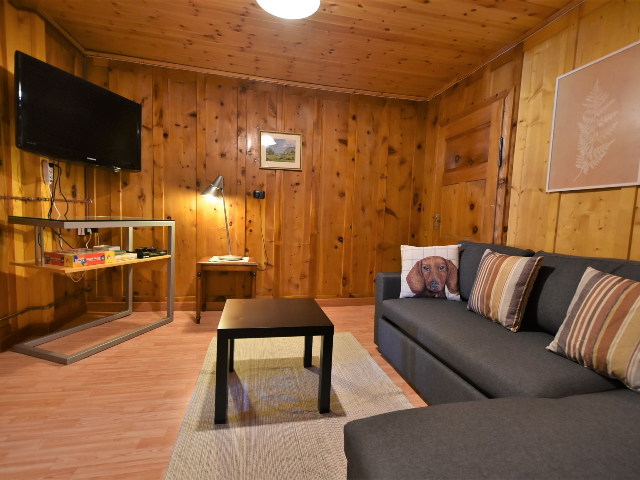 A cozy room with wooden walls, a flat-screen TV mounted on the wall, a sofa with patterned cushions, a small table, and a lamp on a side table.