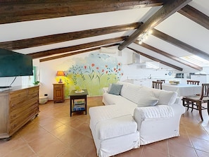 A cozy loft-style living area with a white sofa, wooden beams, a mural, and a kitchen in the background.
