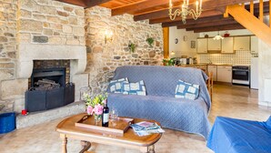 A cozy living room with a stone fireplace, wooden beams, a sofa, and a coffee table with magazines and a bottle. - Avel Vor by Interhome (Saint Pol de Léon)