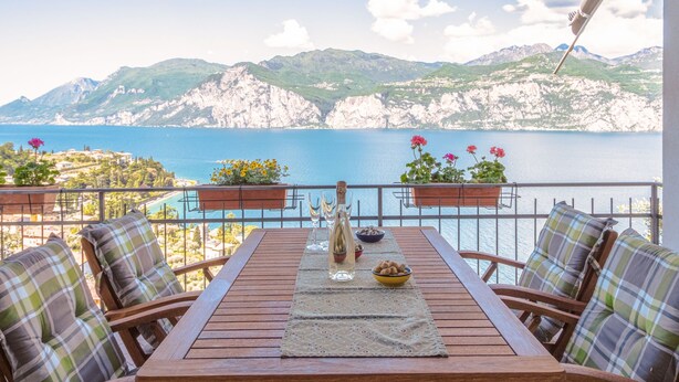 A wooden table with chairs, a bottle, and a bowl of snacks on a balcony overlooking a sea and mountains. - Vacation home Verdin by Interhome (Malcesine)