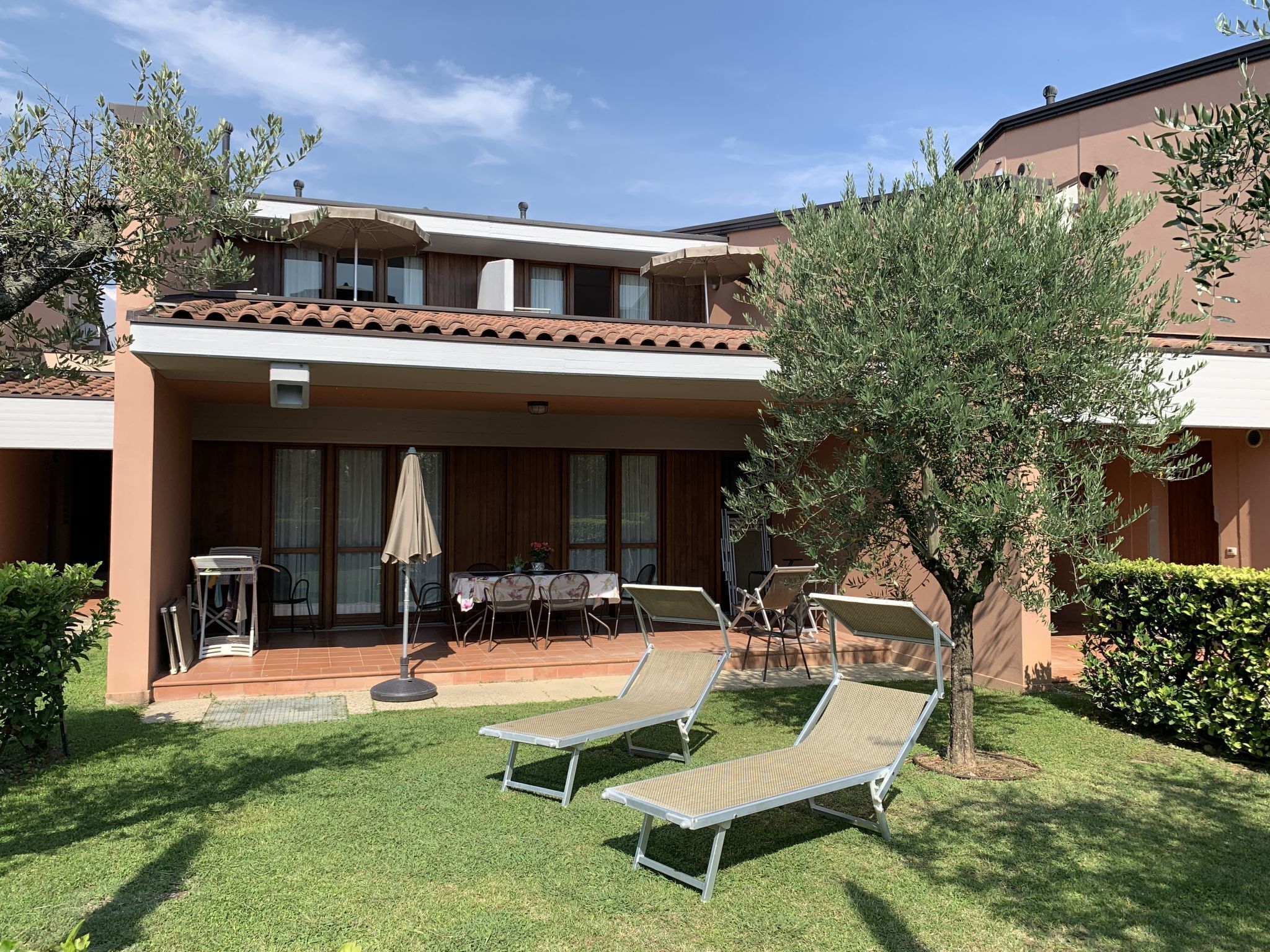 A two-story building with a covered patio area featuring a table and chairs, and two lounge chairs on the lawn.