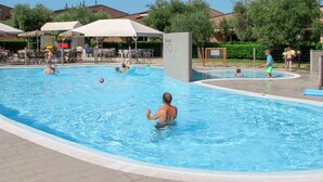 Ein Schwimmbecken mit Schwimmern und spielenden Personen, umgeben von Sonnenschirmen und Liegestühlen.