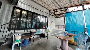 An outdoor patio area with a concrete floor, metal railing, and a blue tarp covering part of the space. There are several tables and chairs, some with wooden tops and others with plastic. A staircase leads to an upper level. - Hotel O Ting Brother Sg Klah Inn (Sungkai)