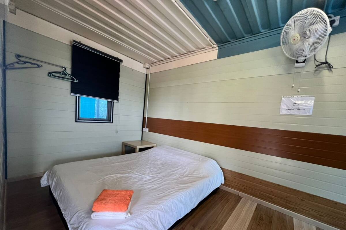 Ein Zimmer mit einem Einzelbett, einem Holzkopfende, einem kleinen Nachttisch, einem Wandventilator und einem Fenster mit Verdunklungsvorhang.