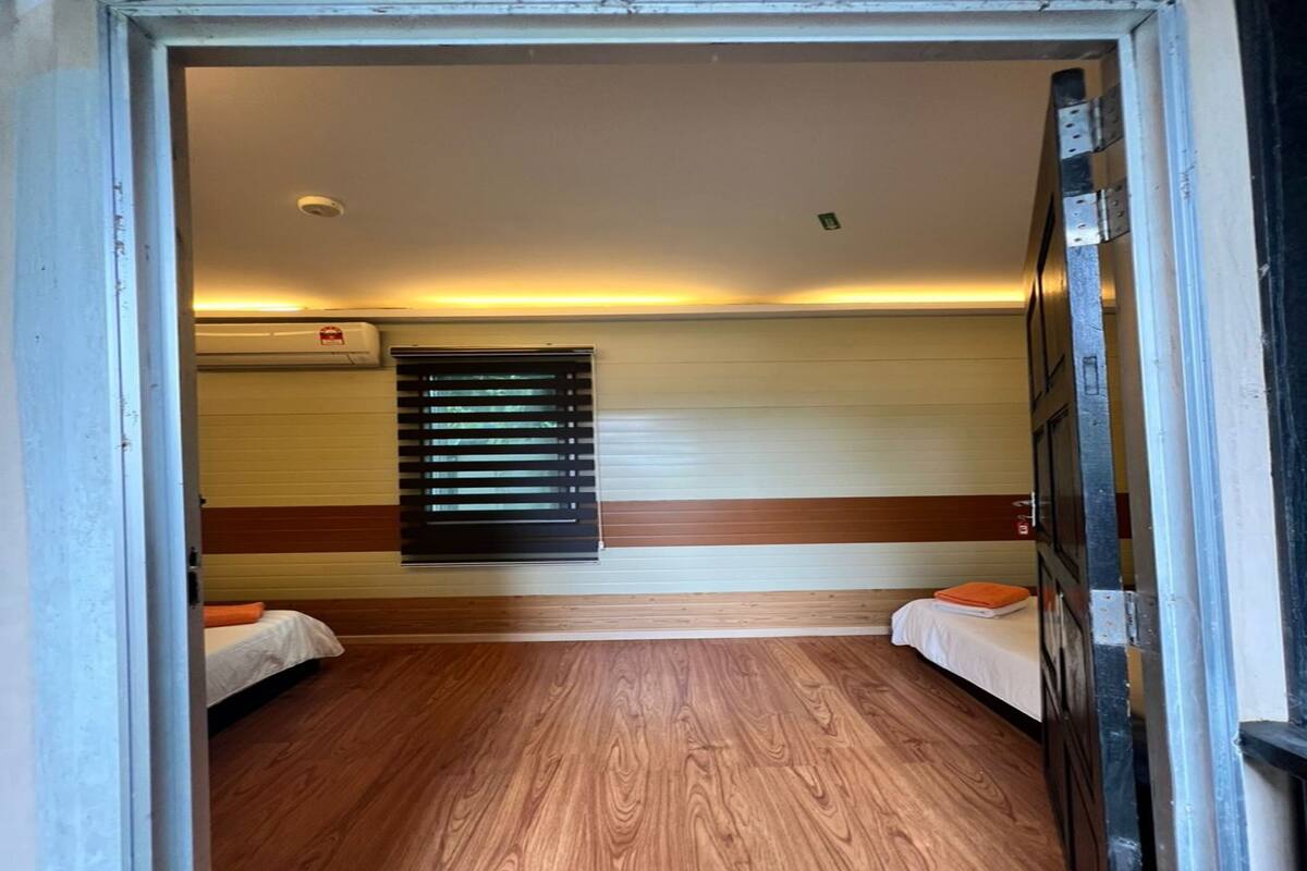 Ein kleines Zimmer mit zwei Betten, Holzboden, einem Fenster mit Jalousien und einer Wand-Klimaanlage.