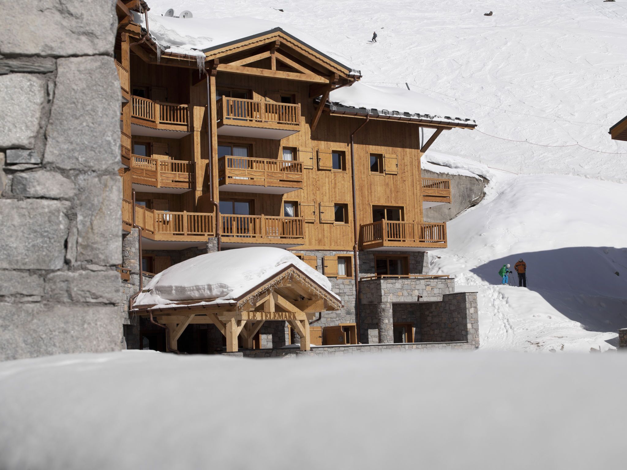 A multi-story wooden chalet with balconies, set against a snowy mountain backdrop.