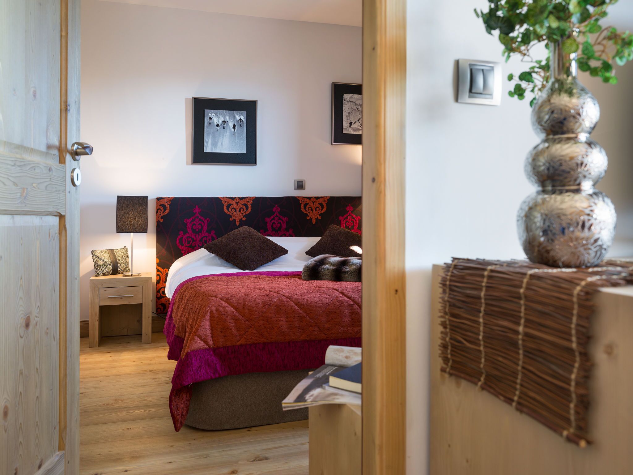 A bedroom with a wooden floor, a bed with a patterned headboard, a bedside table with a lamp, and a wall-mounted picture.
