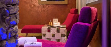 Un cuarto de spa con pared de piedra, techo de madera, una obra de arte enmarcada, camilla de masaje, una silla roja, una mesa de cristal con una vela y una toalla con la etiqueta 'GARITA PARIS'.