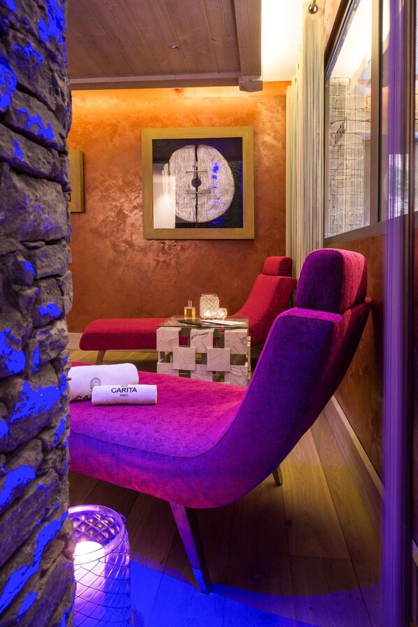 A spa room with a stone wall, a wooden ceiling, a framed artwork, a massage table, a red chair, a glass table with a candle, and a towel labeled "GARITA PARIS".