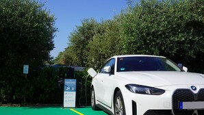 Ein weißer BMW parkt an einer Ladestation für Elektrofahrzeuge mit einem grünen Parkplatz und Bäumen im Hintergrund.