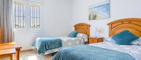 A bedroom with two beds, a wooden headboard, a nightstand, a table, and a window with bars.