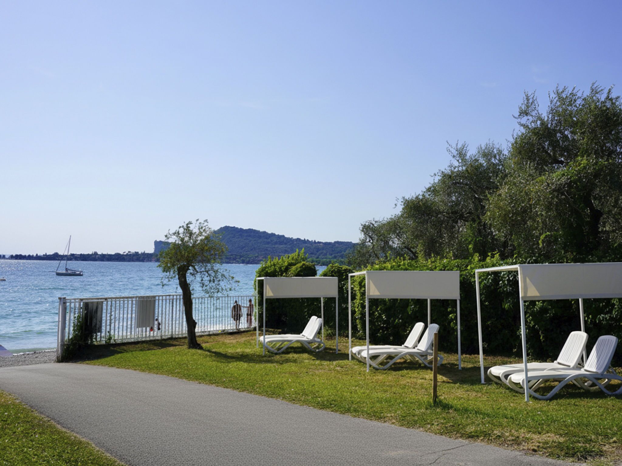 Un espace aménagé au bord du lac, avec des chaises longues sous une pergola, un sentier pédestre et une vue lointaine sur un voilier.