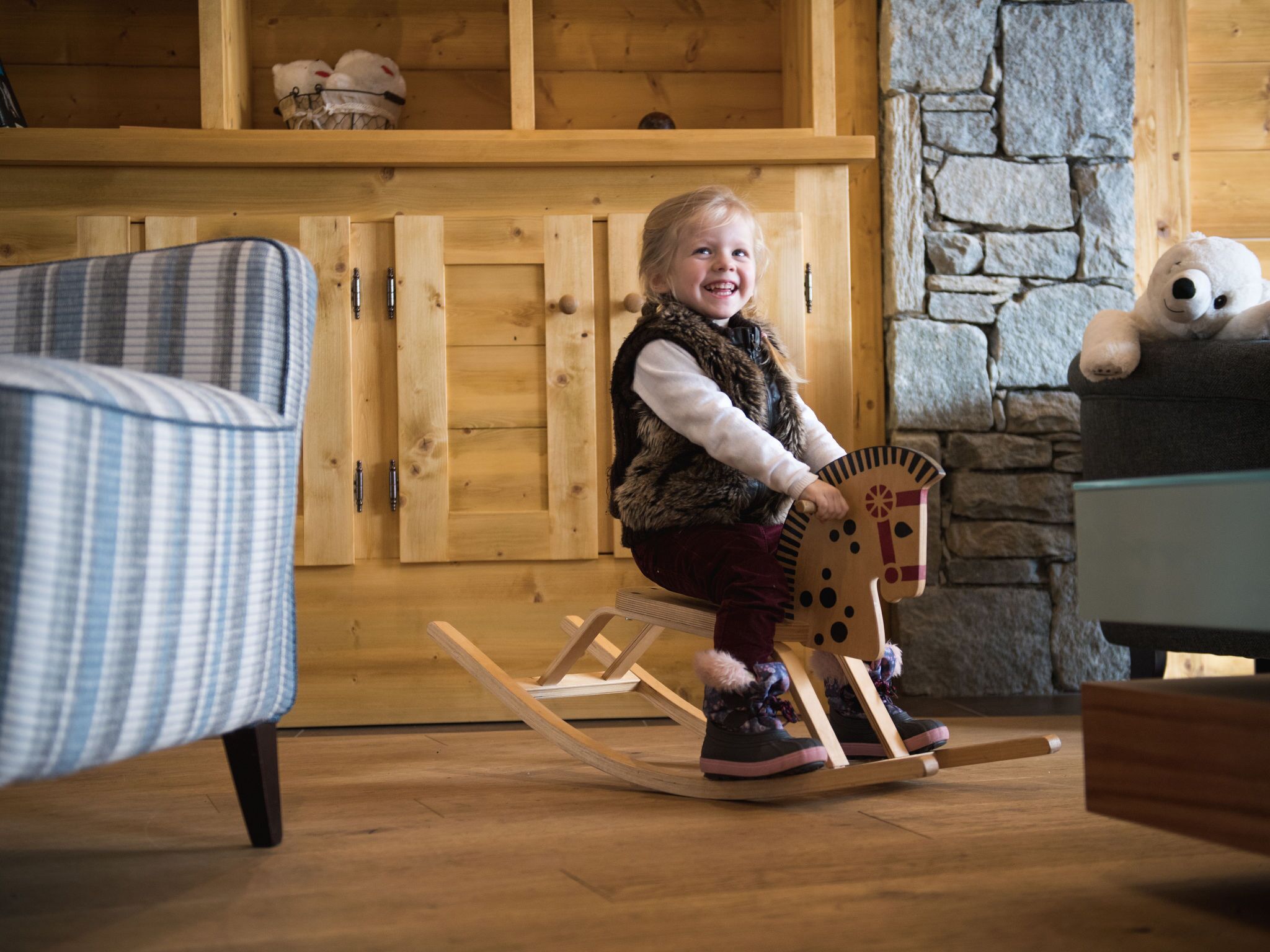A child on a wooden rocking horse in a room with wooden furniture and a stone wall.