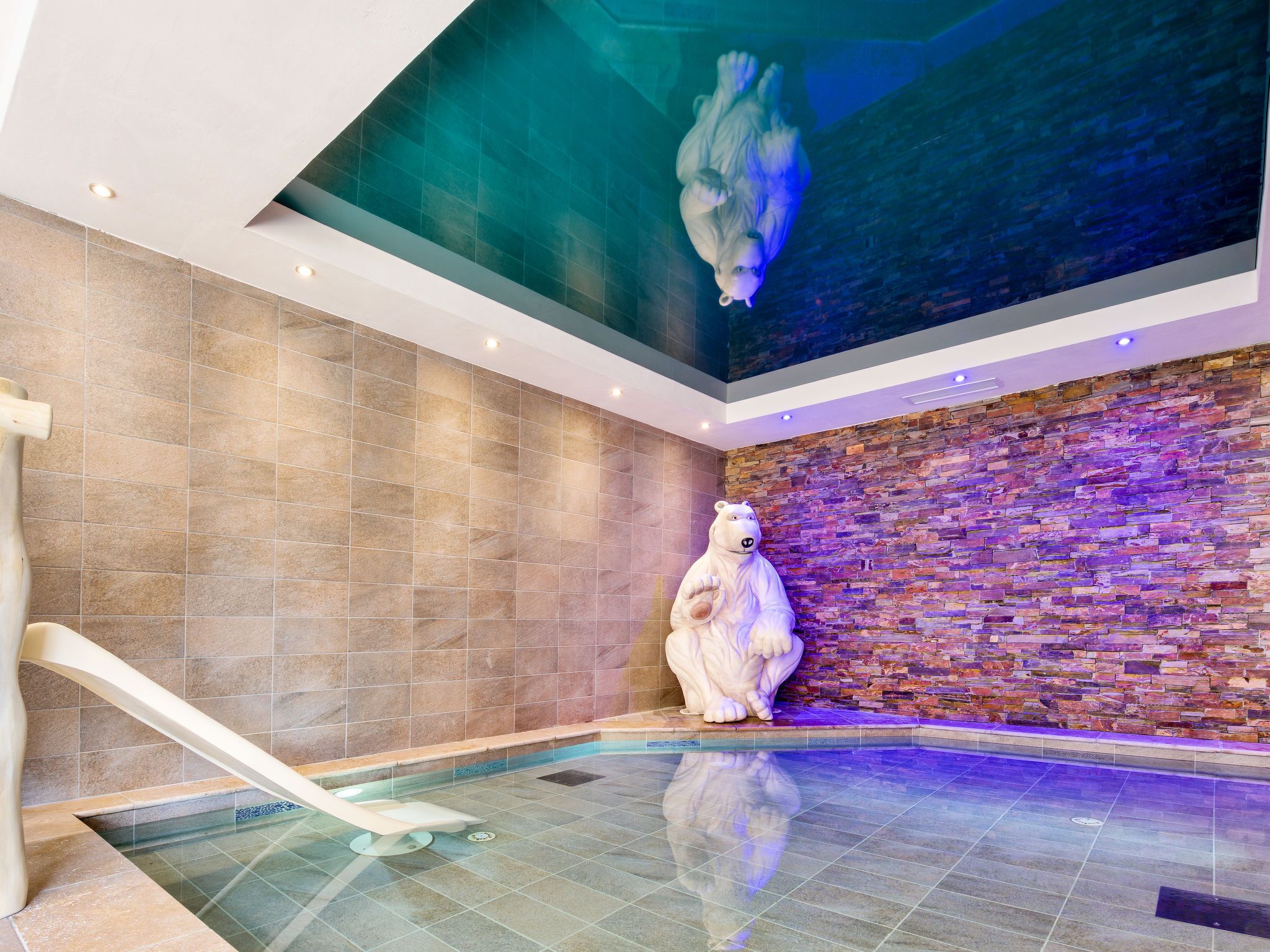 A modern indoor pool area with a large bear statue, a stone wall, and a glass ceiling.