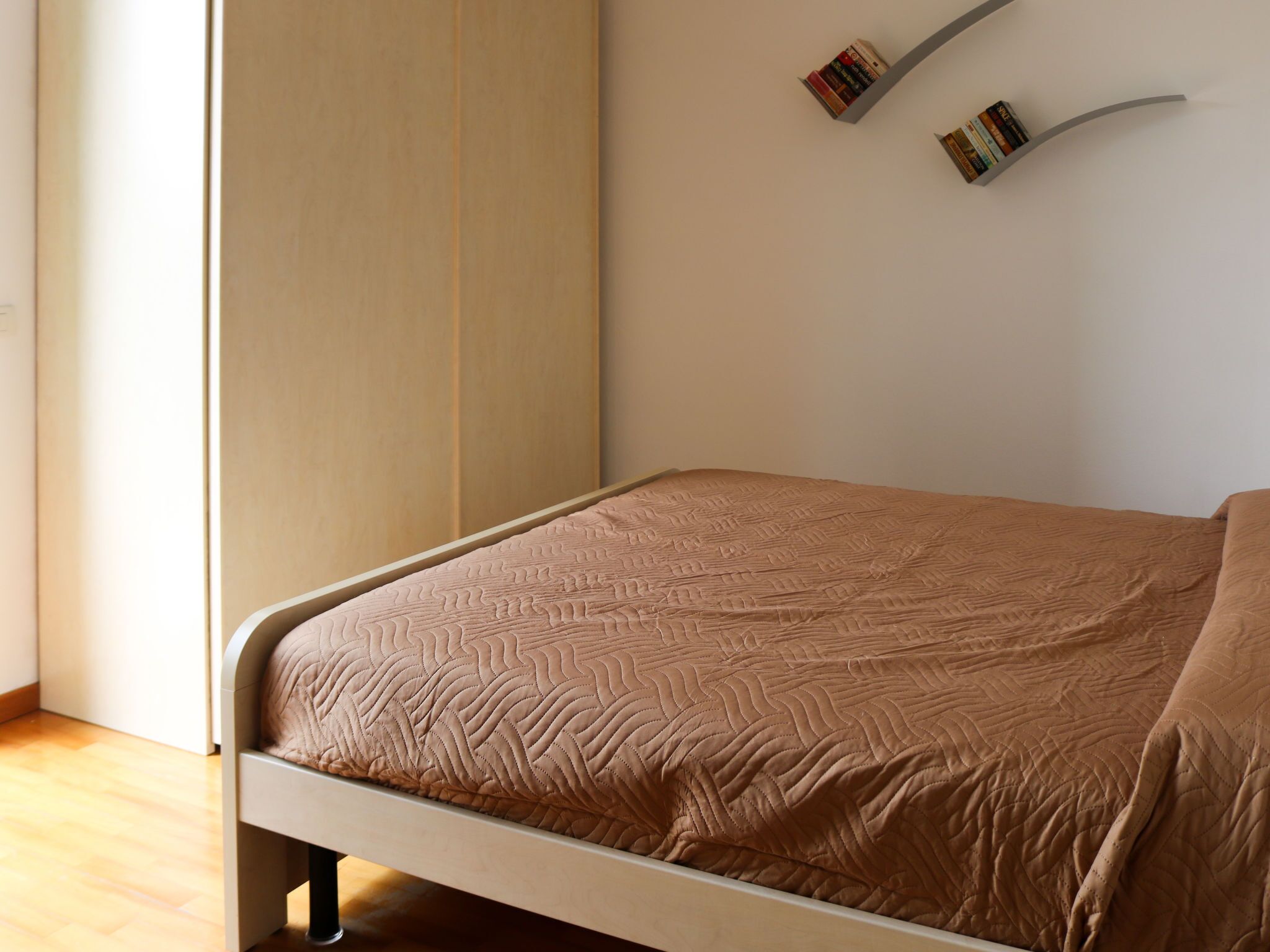 Un dormitorio con cama, suelo de madera, estantería empotrada con libros y una ventana.