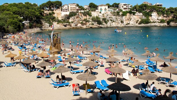 A beach with numerous sun loungers and umbrellas, a lifeguard tower, and a clear view of the coastline with buildings.