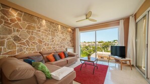 A living room with a stone wall, a brown sofa with colorful cushions, a red rug, a TV on a stand, and a large window with a view of buildings and greenery.