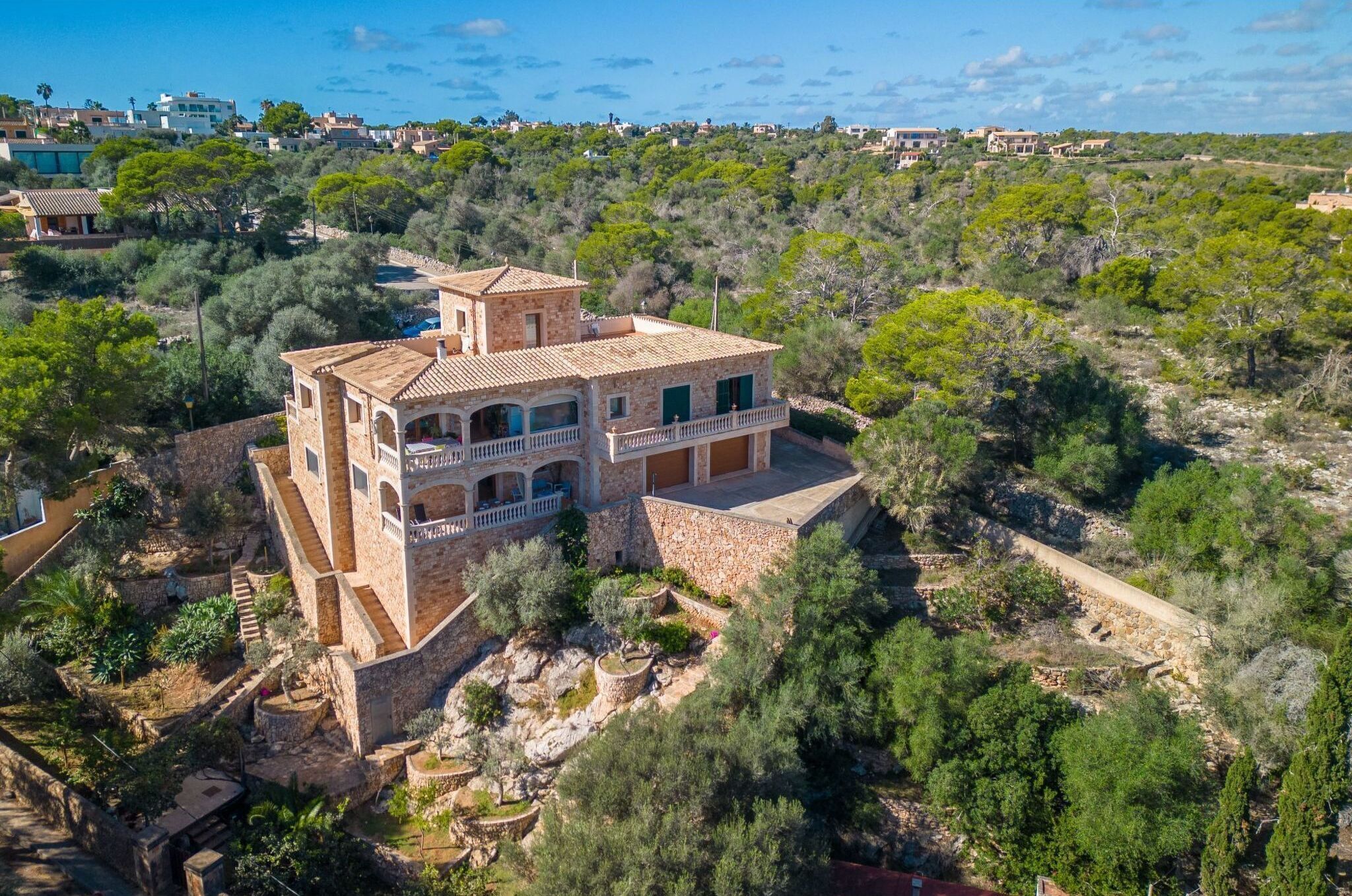 Une grande maison en pierre à plusieurs étages, dotée d’un balcon, entourée d’une végétation luxuriante et située dans un paysage vallonné.