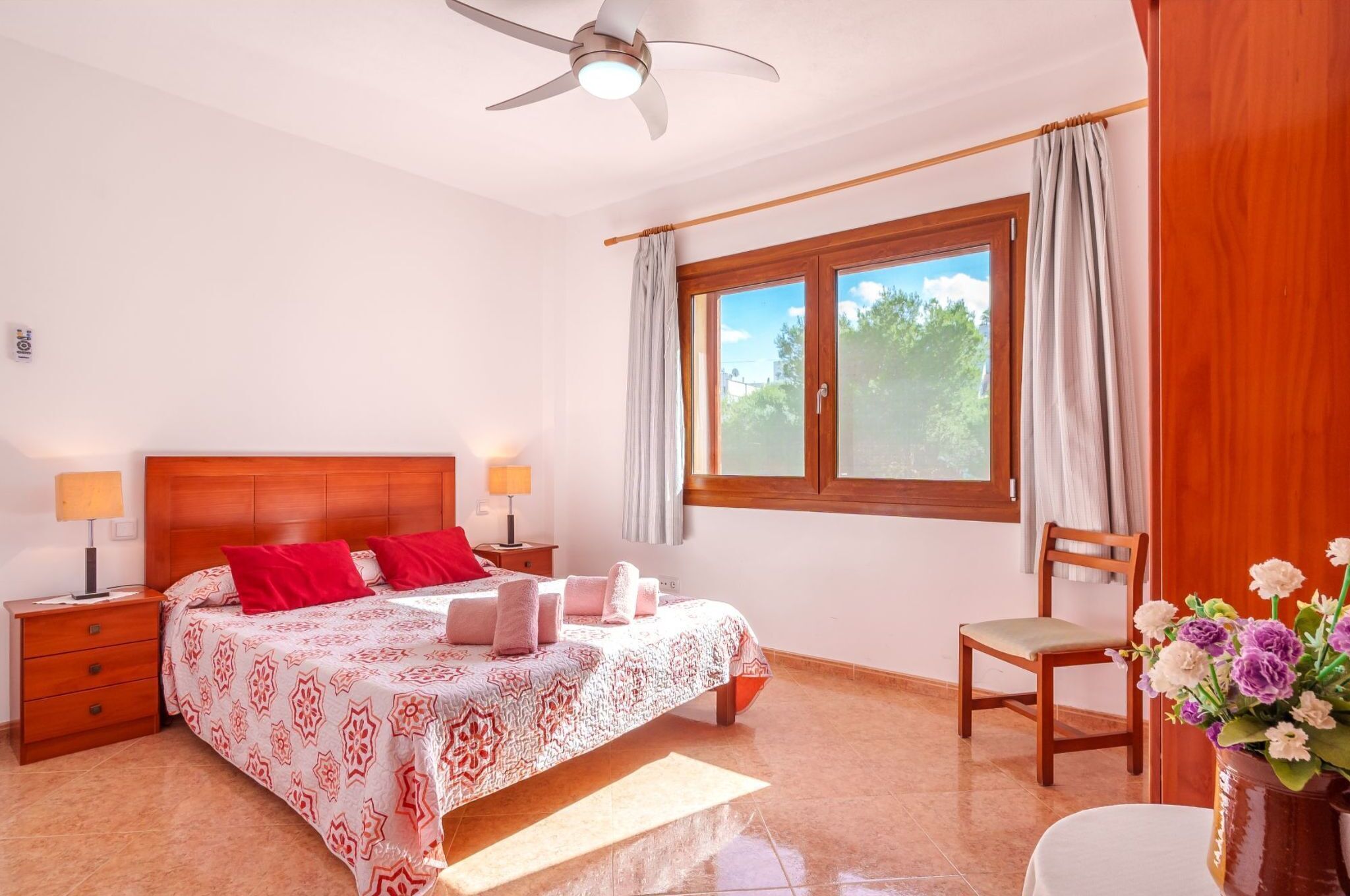 Une chambre à coucher comprenant un lit, deux tables de chevet, une chaise, une fenêtre avec des rideaux, un ventilateur de plafond et un vase avec des fleurs.