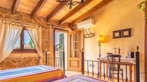 Ein gemütliches Schlafzimmer mit Steinwand, Holzbalken, einer farbenfrohen gesteppten Bettdecke, einem Schreibtisch mit Lampe und Blick durch ein Fenster mit Vorhängen.