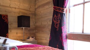 Une pièce chaleureuse avec un décor intérieur en bois, un lit recouvert d’une couverture rose, une table de chevet avec une tasse et un bol de collations, et une fenêtre avec des rideaux.