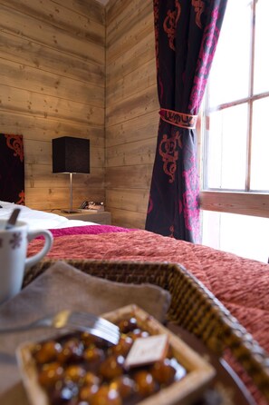 Een knus ingerichte kamer met houten details, een bed met een roze dekbed, een nachtkastje met een kopje en een schaaltje lekkernijen, en een raam met gordijnen.